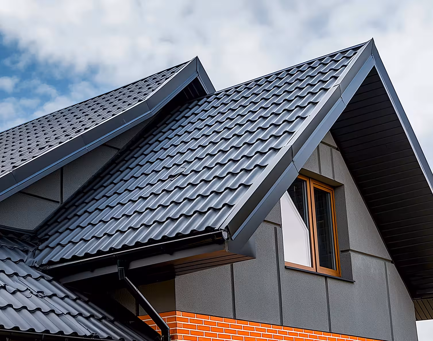 Close-up of a modern house roof with dark metal tiles and a window under a cloudy sky.