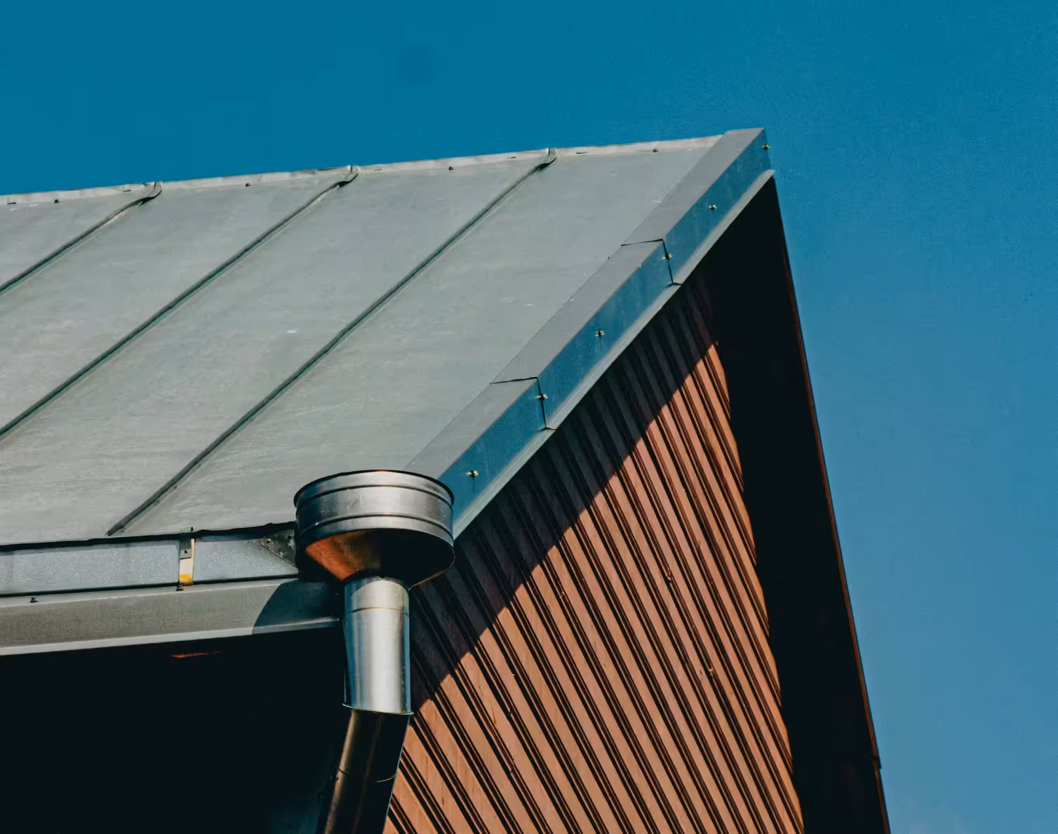 Close-up of a metal roof with a gutter and a vent pipe against a clear blue sky.