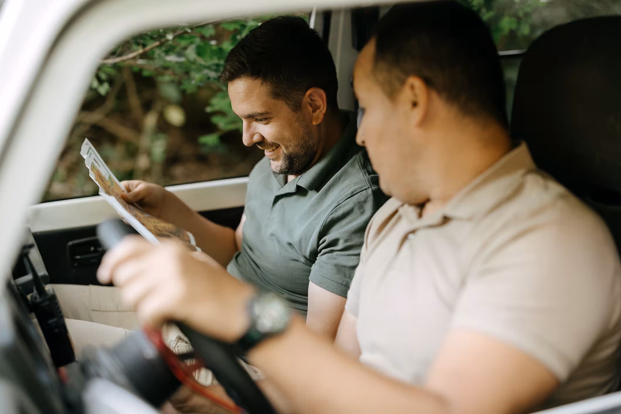 Two men inside a vehicle looking at a map, one holding the steering wheel, outdoors visible.