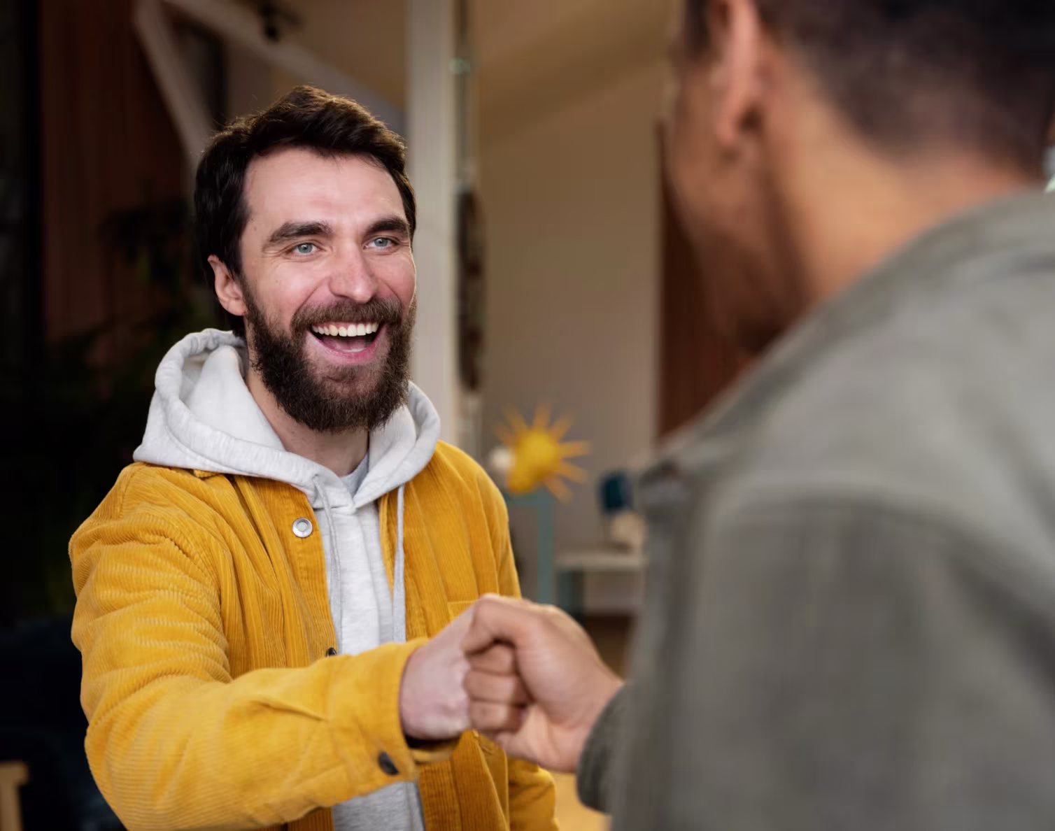 Two men fist bumping with one smiling and wearing a yellow jacket indoors.