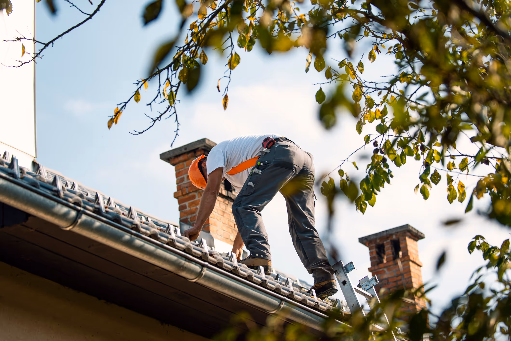 Construction worker in orange helmet fixing roof tiles near brick chimneys during daytime.
