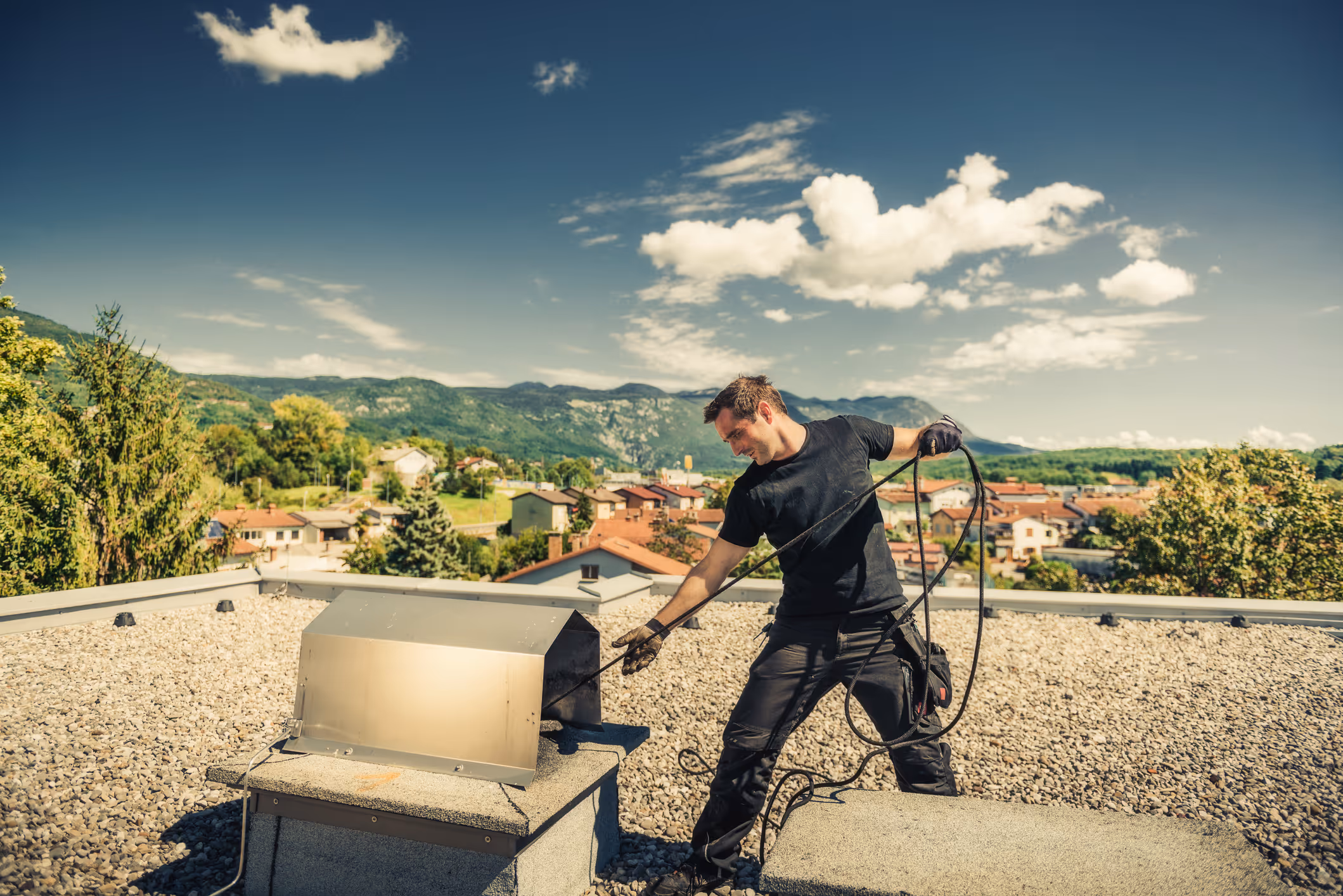 Man in black shirt cleaning chimney on gravel roof with scenic town and mountains in background.