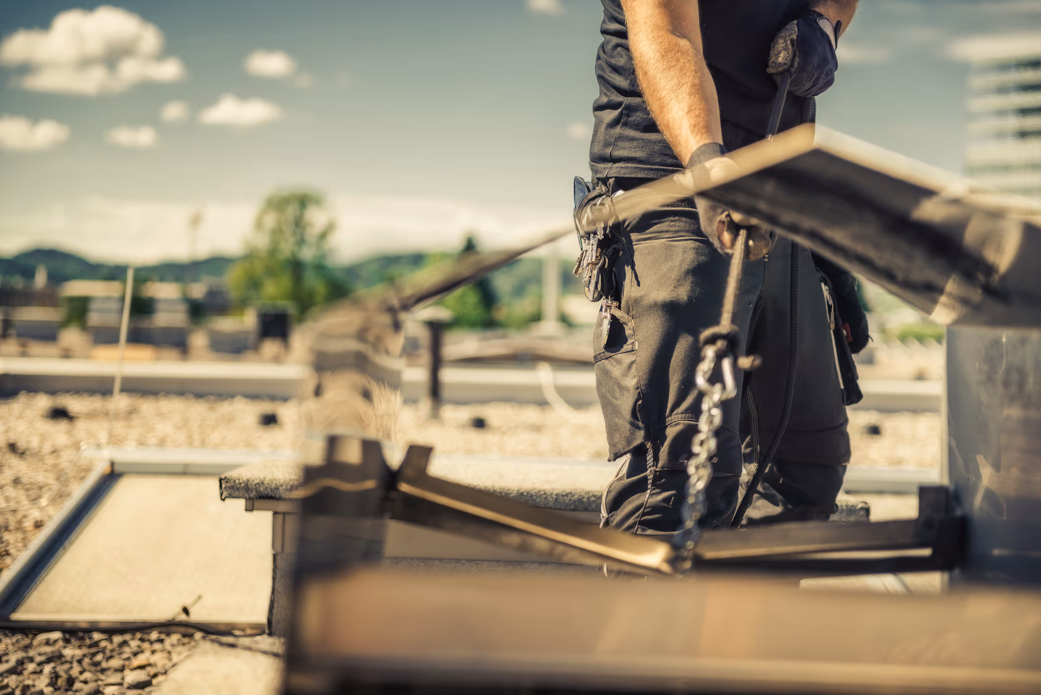 Roofer in work pants and gloves securing equipment on a rooftop with safety harness chains.