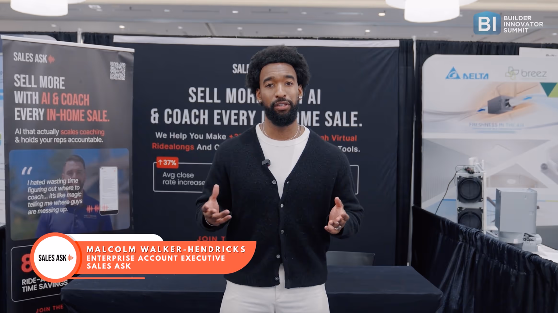 Man in black cardigan and white shirt speaking at a sales booth with promotional banners behind him.