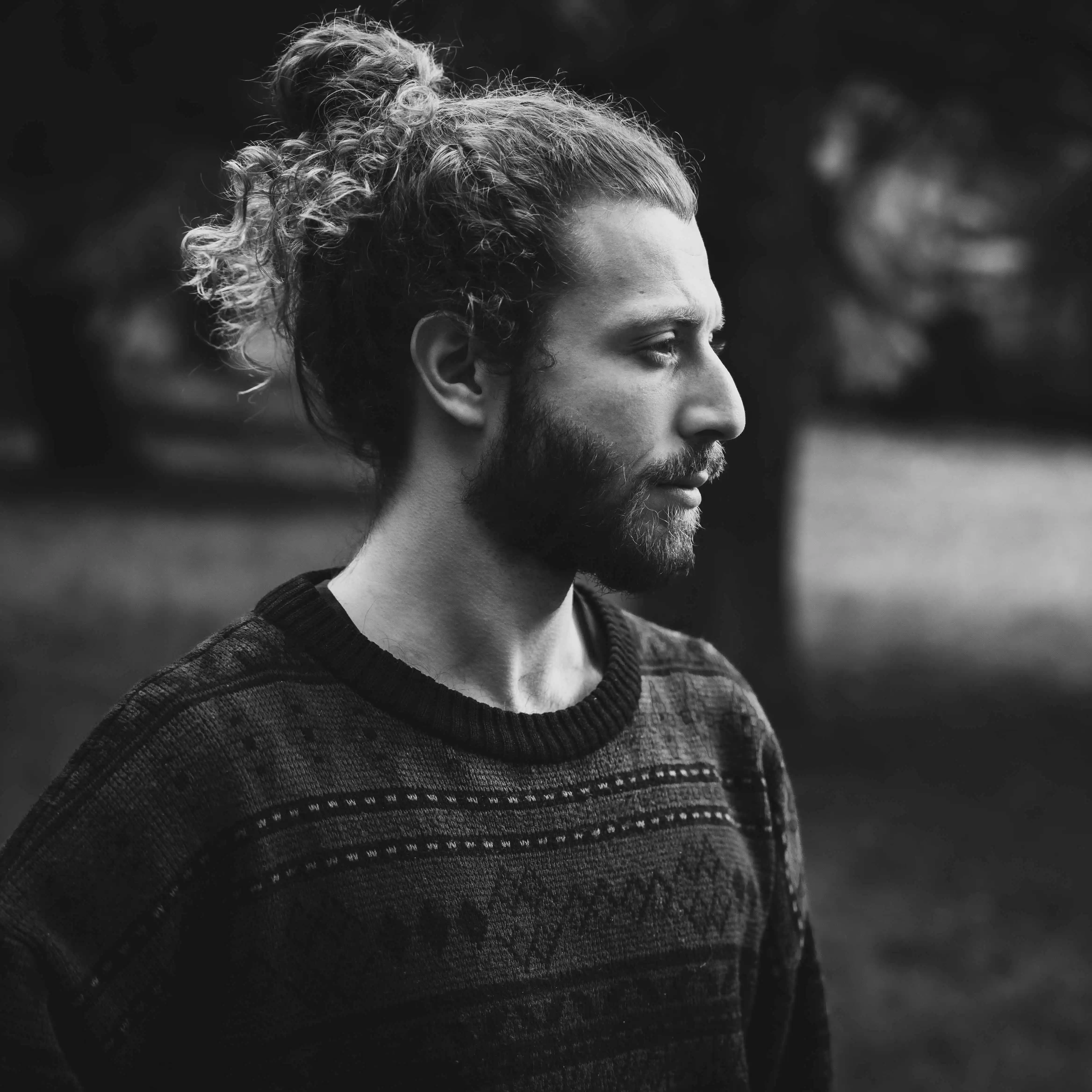 Portrait en noir et blanc d'un homme barbu aux cheveux bouclés attachés, regardant vers la droite.