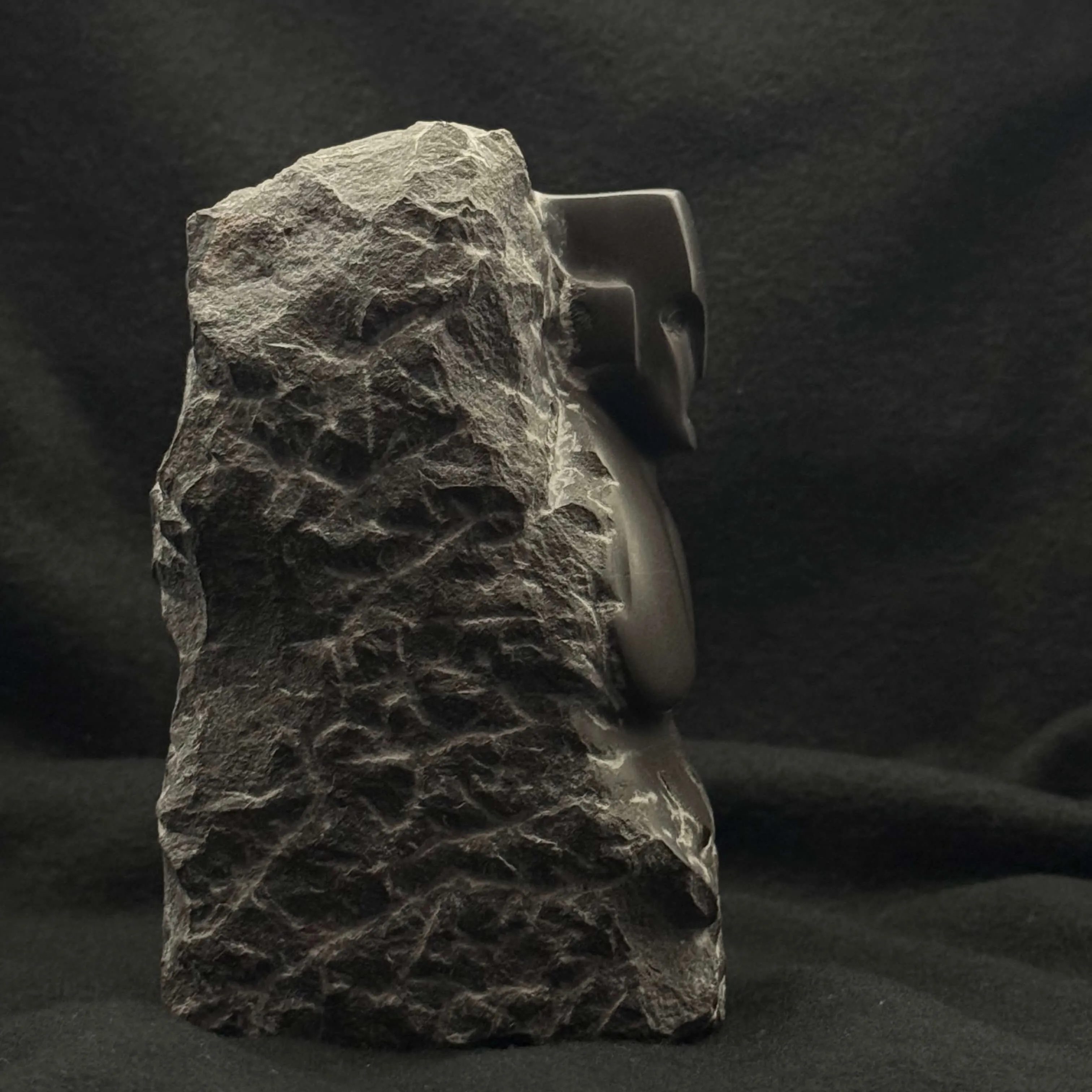 Sculpture of a face partly emerging from rough stone against a dark background.