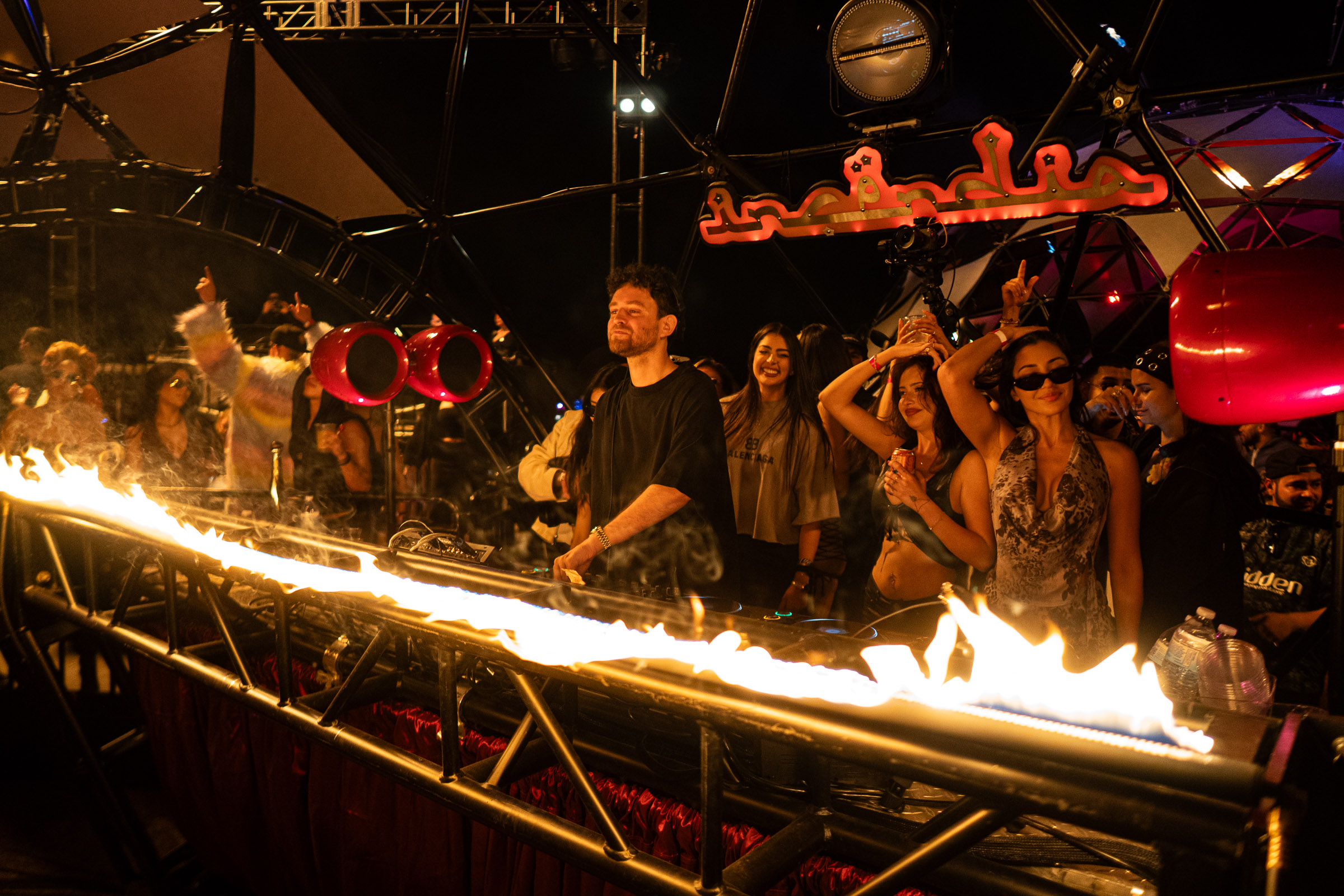 DJ performing at night with crowd enjoying nearby and flames in front of the stage.