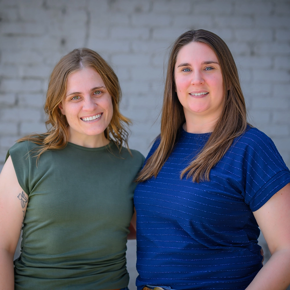 Becca and Jenn smiling standing close together in front of a gray brick wall.