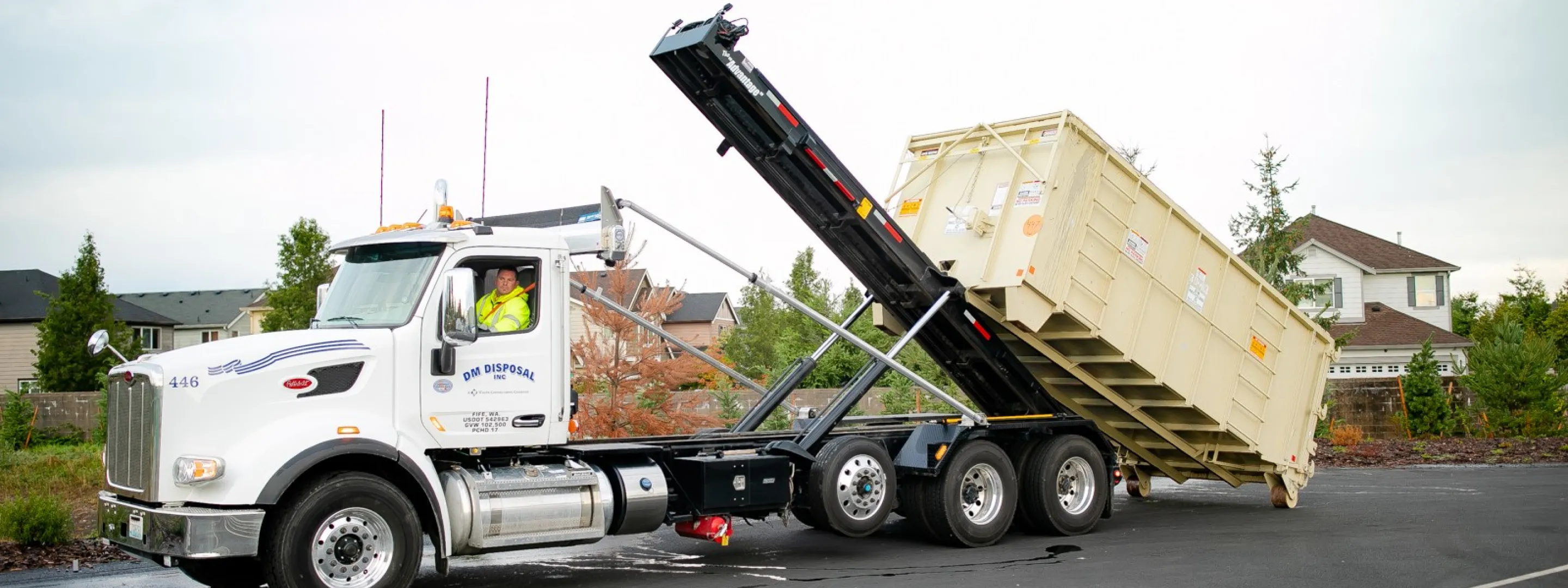 White roll-off truck lifting a large beige dumpster container in a suburban neighborhood.