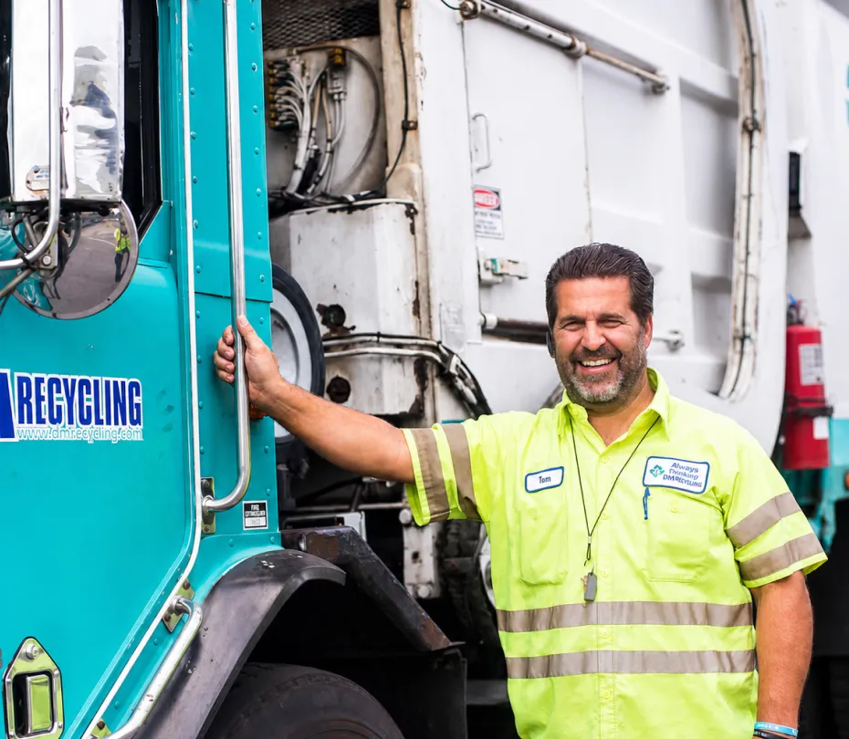 Smiling recycling worker named Tom in a neon safety shirt standing next to a blue recycling truck.