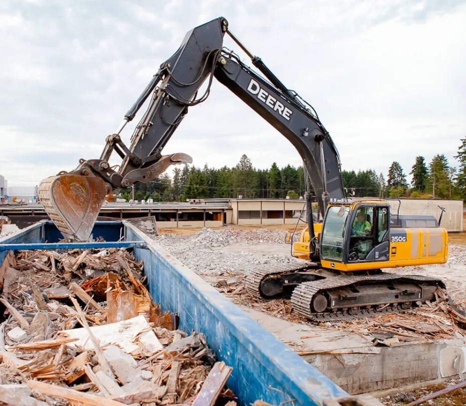 John Deere excavator loading debris into a large blue container at a recycling site.