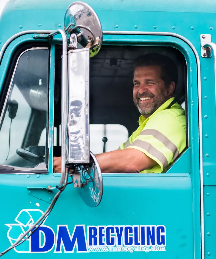 Smiling man in a yellow safety shirt sitting in a teal DM Recycling truck cab.