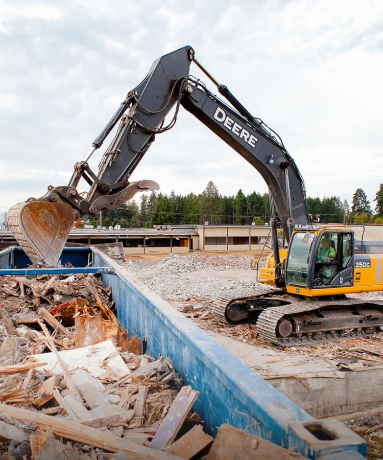 Excavator loading construction debris into a large blue recycling bin at a demolition site.