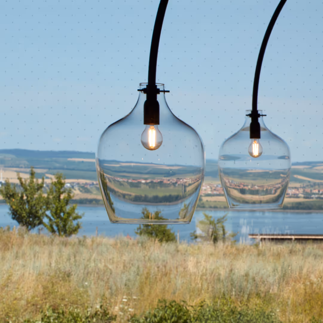 Dvě průhledné závěsné lampy se žárovkami na pozadí louky, řeky a vzdálených kopců za jasného dne.