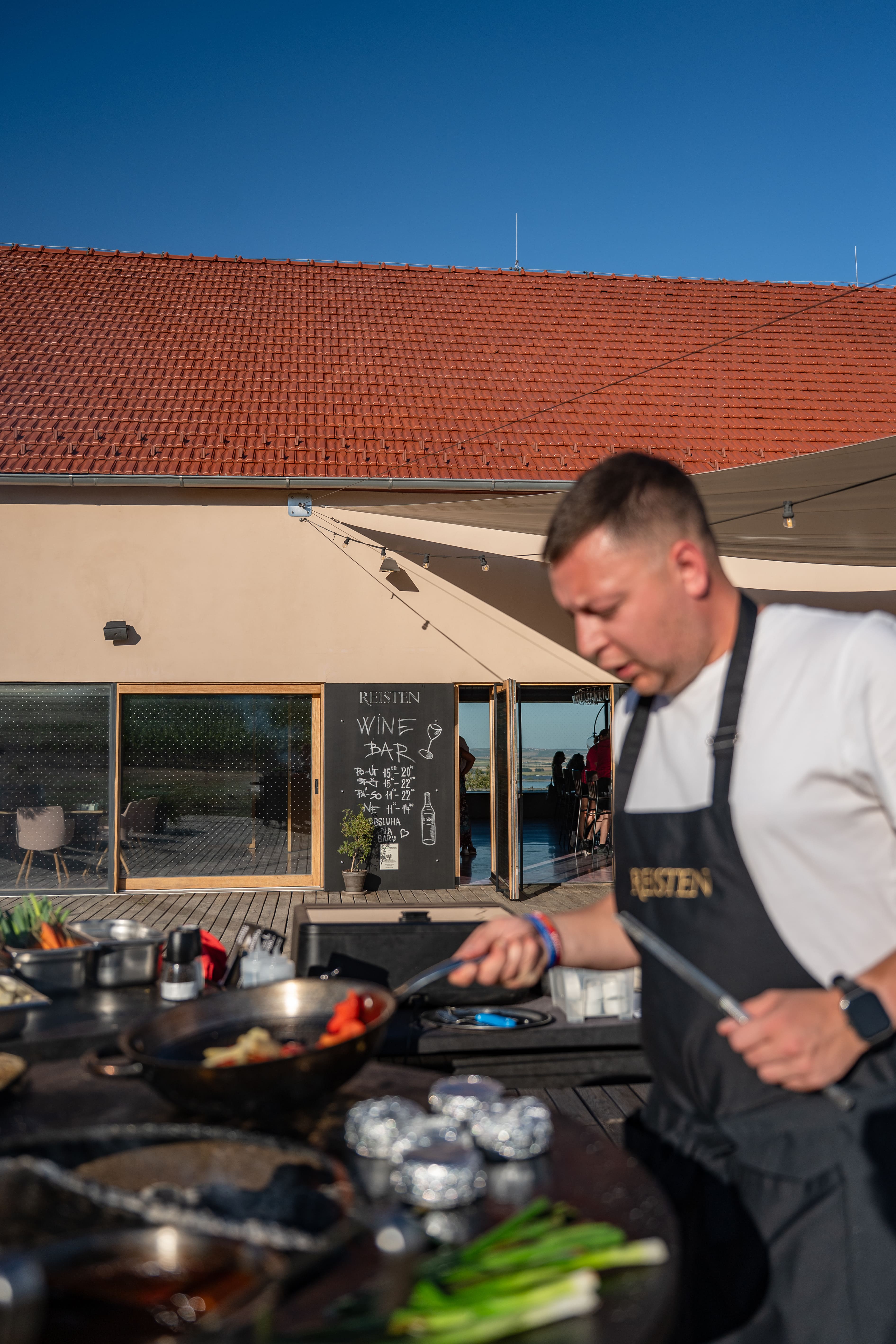 Muž v zástěře připravuje jídlo u venkovního grilu s nápisem Reisten Wine Bar na pozadí.