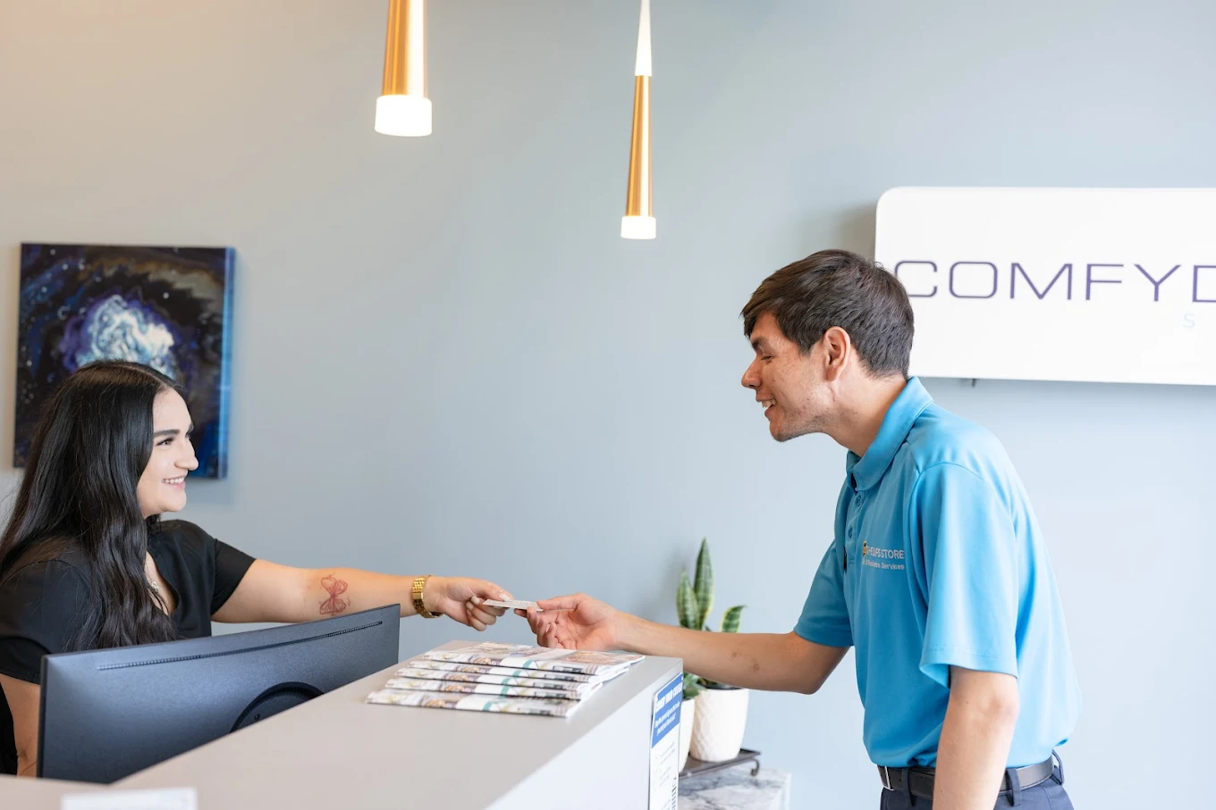 Friendly Comfydent front desk receptionist handing paperwork to a smiling patient at the check-in counter with pendant lighting and Comfydent branding visible