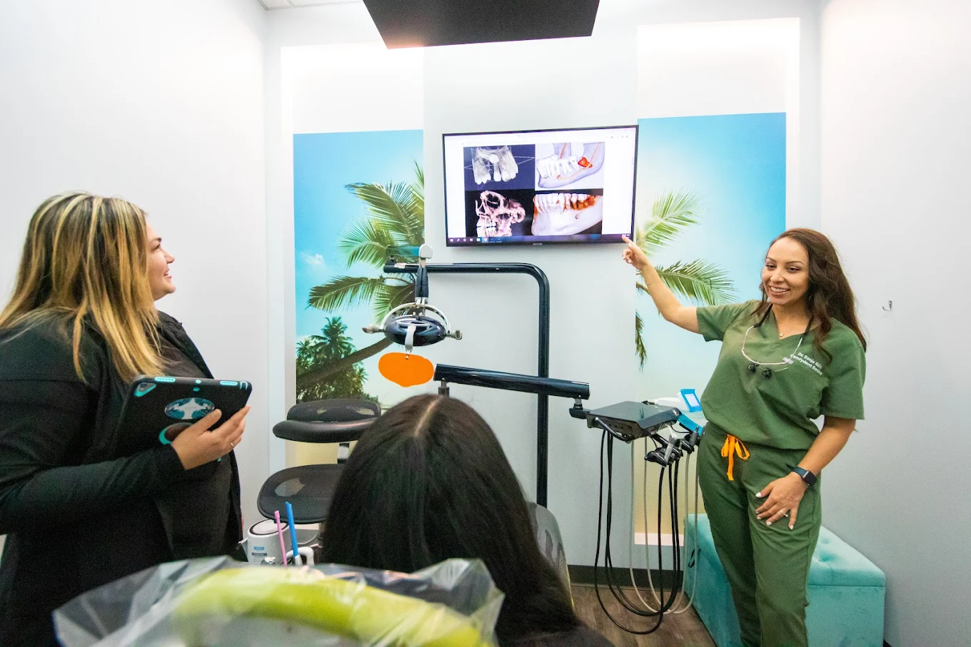 Dental team member in green scrubs reviewing treatment images on a monitor with two patients in a treatment room