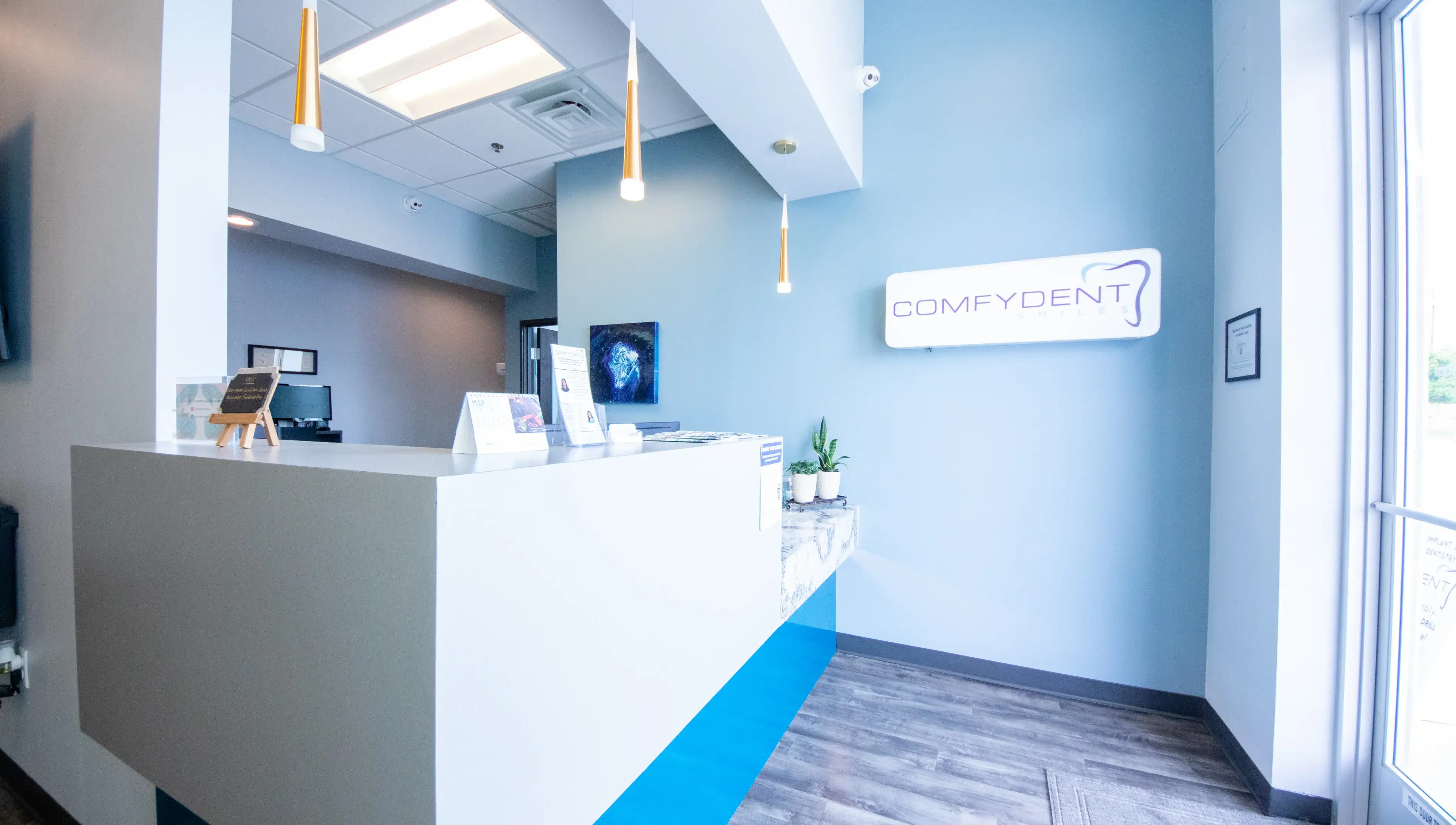 Clean and modern Comfydent dental office reception desk with bright pendant lighting, a white front counter with blue accent, and the Comfydent logo on the wall