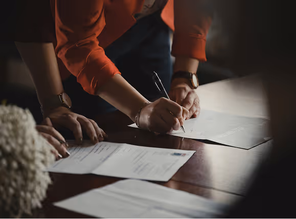 Two people leaning over a table with papers, one person signing a document with a pen.