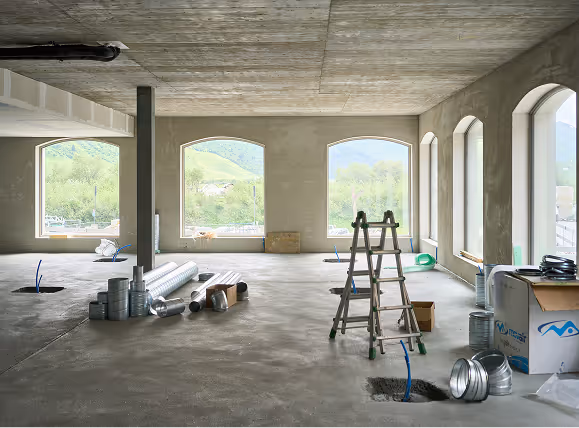 Interior of a building under construction with large arched windows, ladder, and scattered duct pipes.