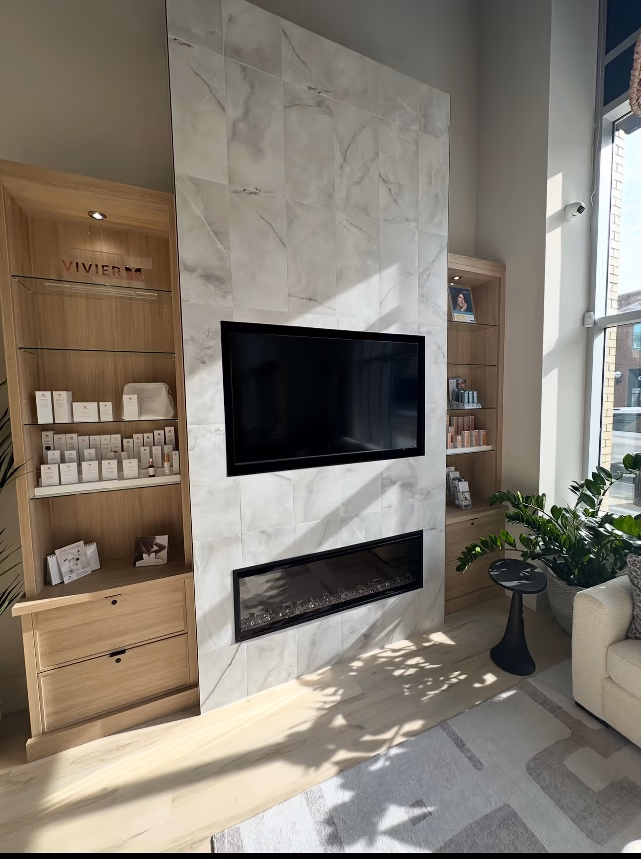 Modern living room corner with a wall-mounted TV above a sleek electric fireplace and wooden shelves with skincare products.