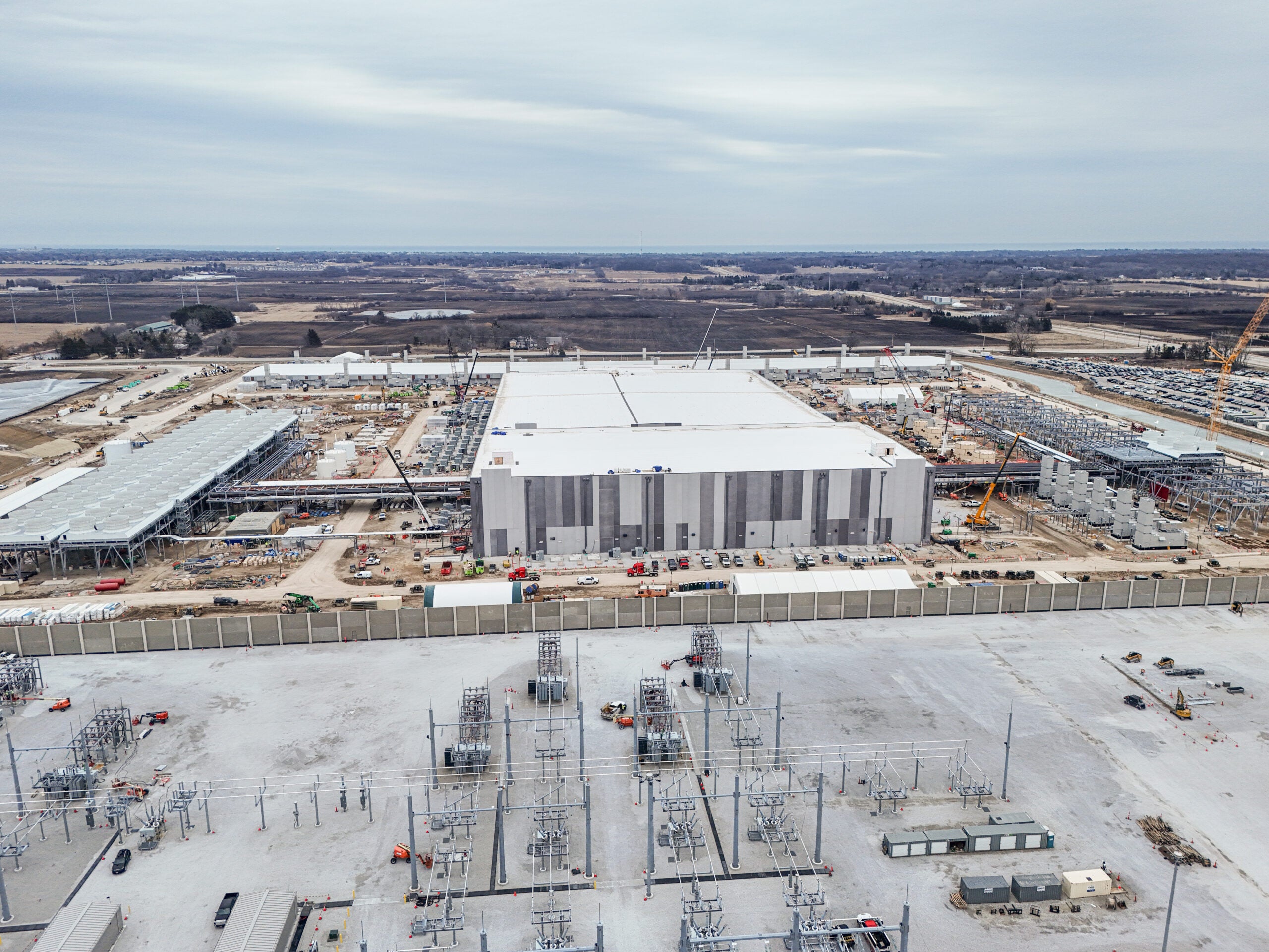 Aerial view of a large industrial building under construction, surrounded by cranes and construction equipment, with fields in the background.