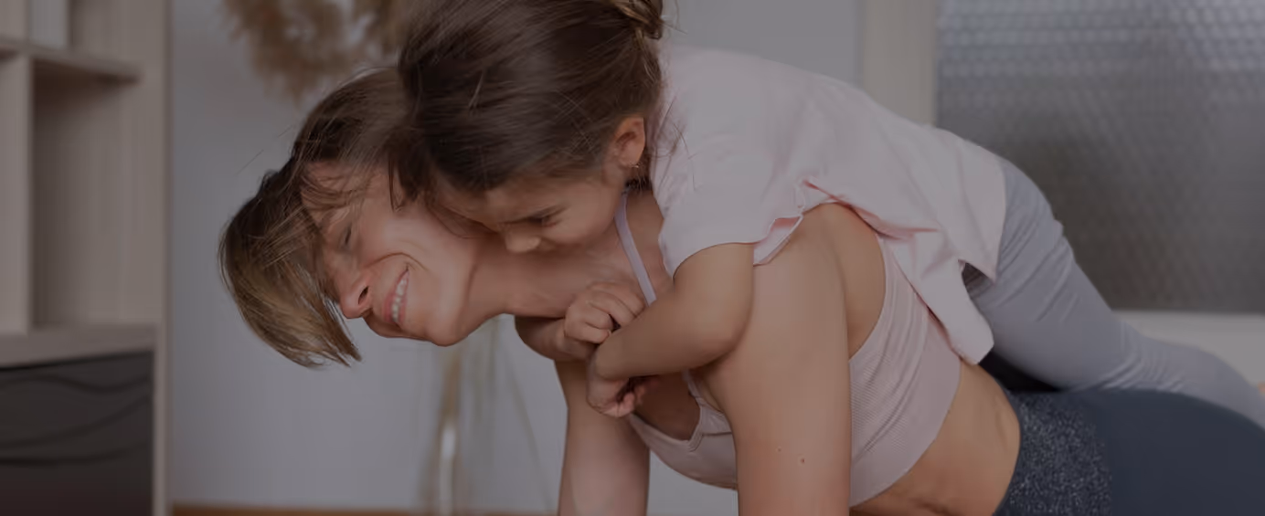 Woman doing a plank exercise with a child playfully on her back in a home setting.