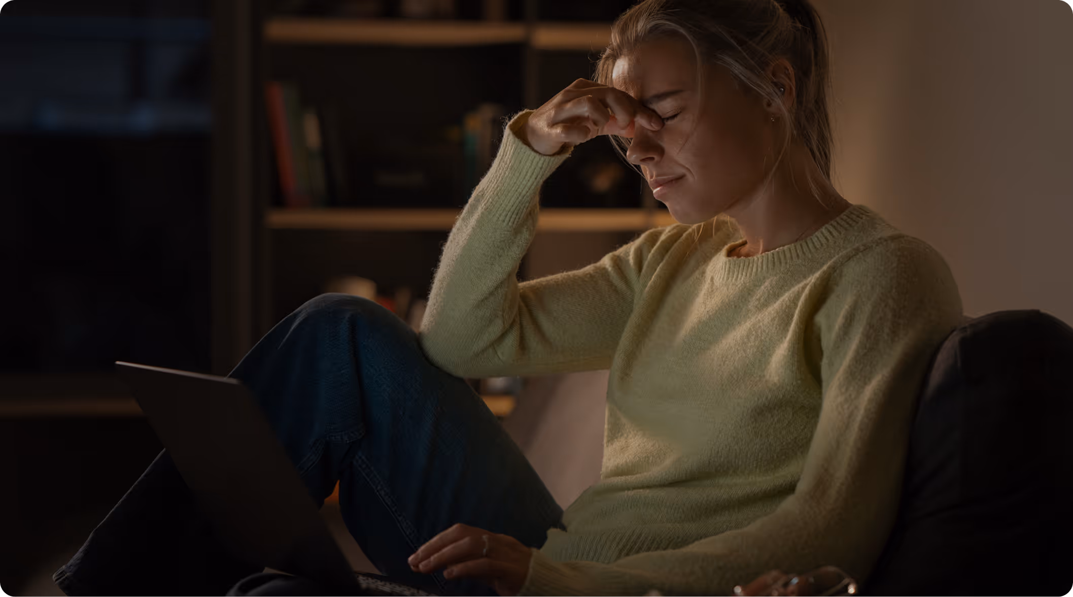 Woman sitting on couch with laptop, pinching the bridge of her nose and eyes closed, looking stressed.