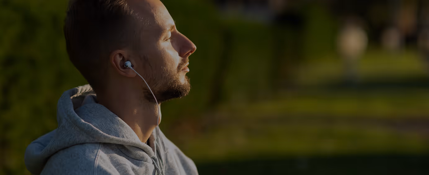 Young man in hoodie outdoors with eyes closed, listening to music on wired earbuds with sunlight on face.