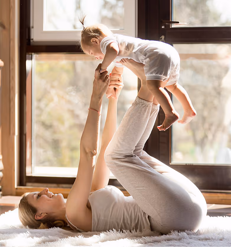 Woman lying on back lifting baby above her with legs and hands inside a bright room.