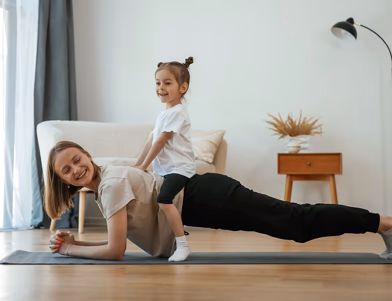 Woman doing plank exercise on a mat with a smiling child sitting on her back in a living room.