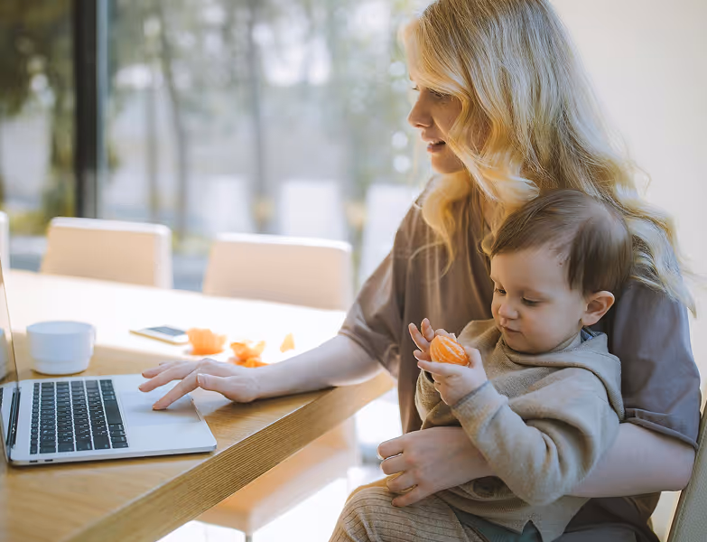 Woman holding toddler while using a laptop at a table with peeled orange slices nearby.