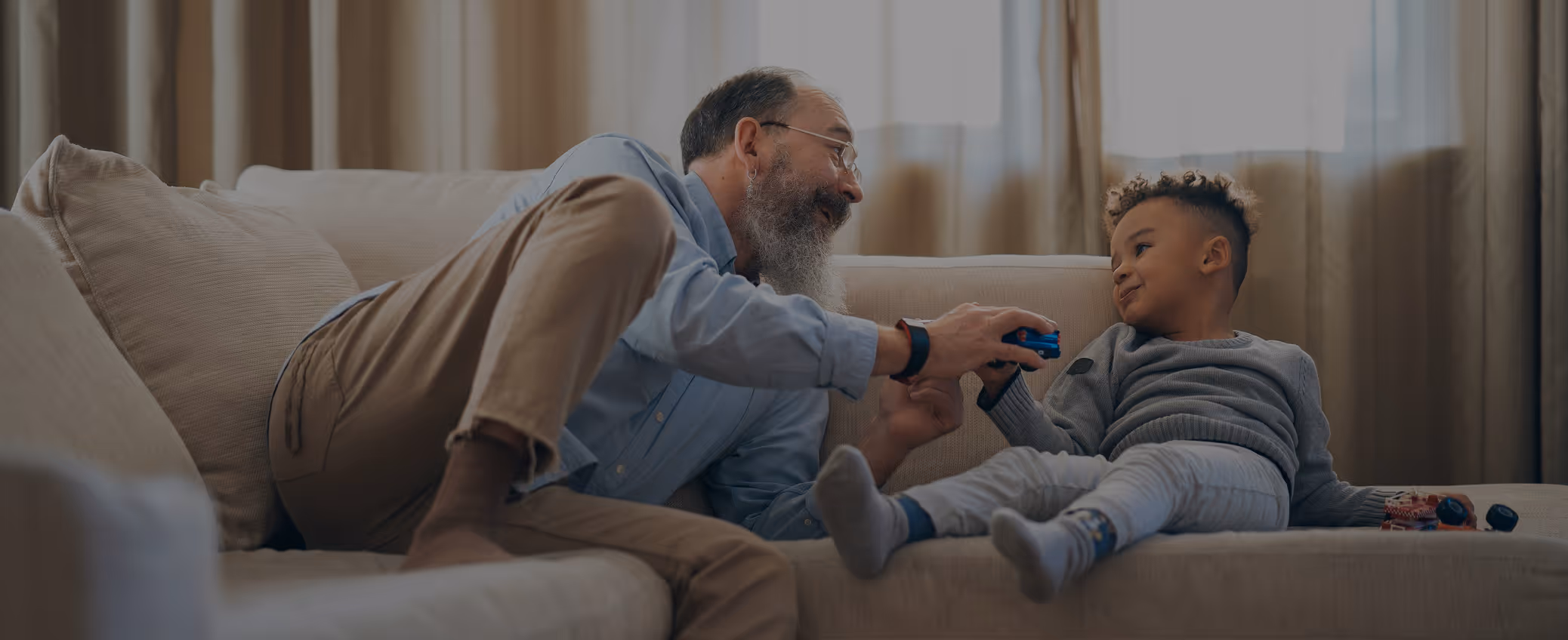 Older man and young boy playing with toy cars together on a beige couch at home.