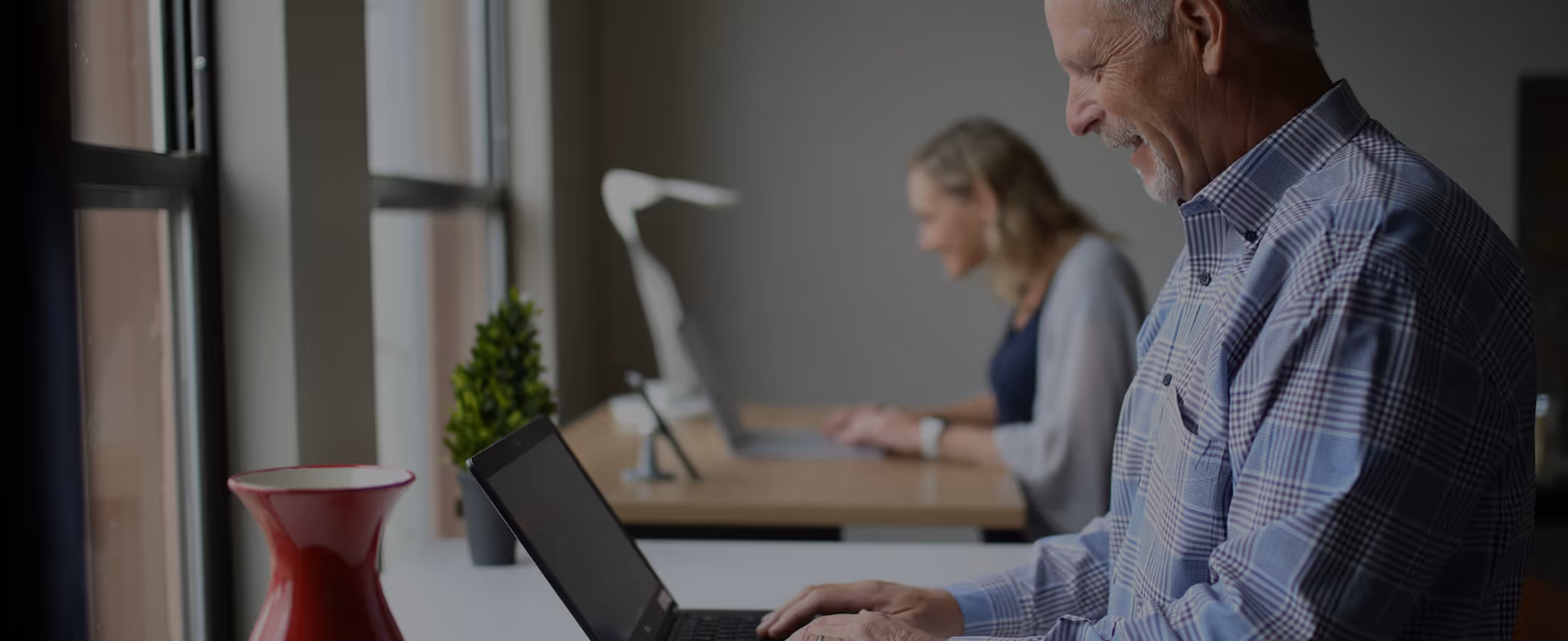 Two people working on laptops in a bright office near a window with a red vase and green plant nearby.