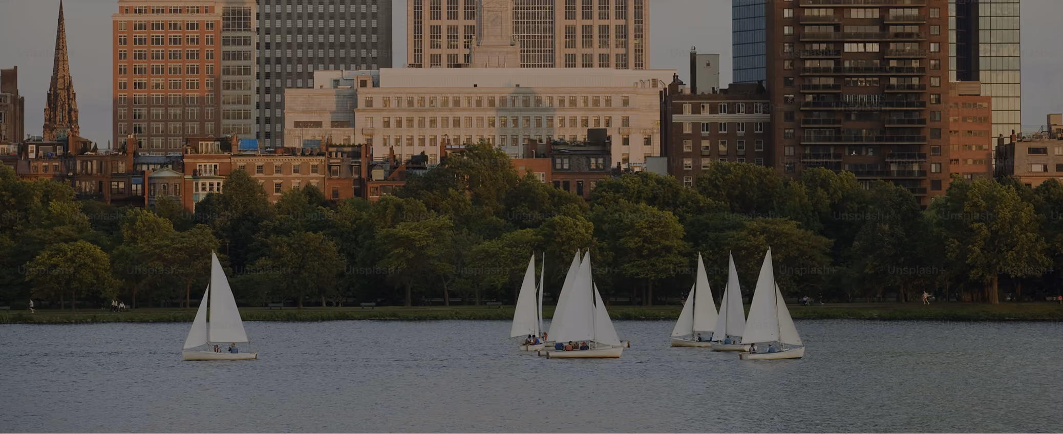 Sailboats on the Charles River with Boston skyline and trees in the background at sunset.