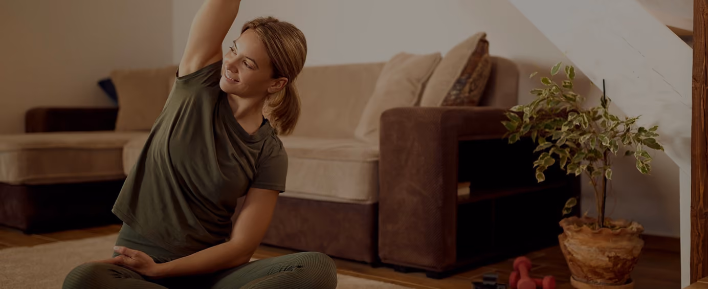 Smiling woman in green clothes stretching arm while sitting cross-legged on floor at home.