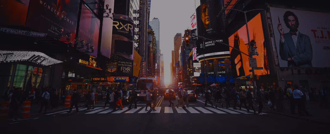 Times square crosswalk at sunset with numerous illuminated billboards and pedestrians.