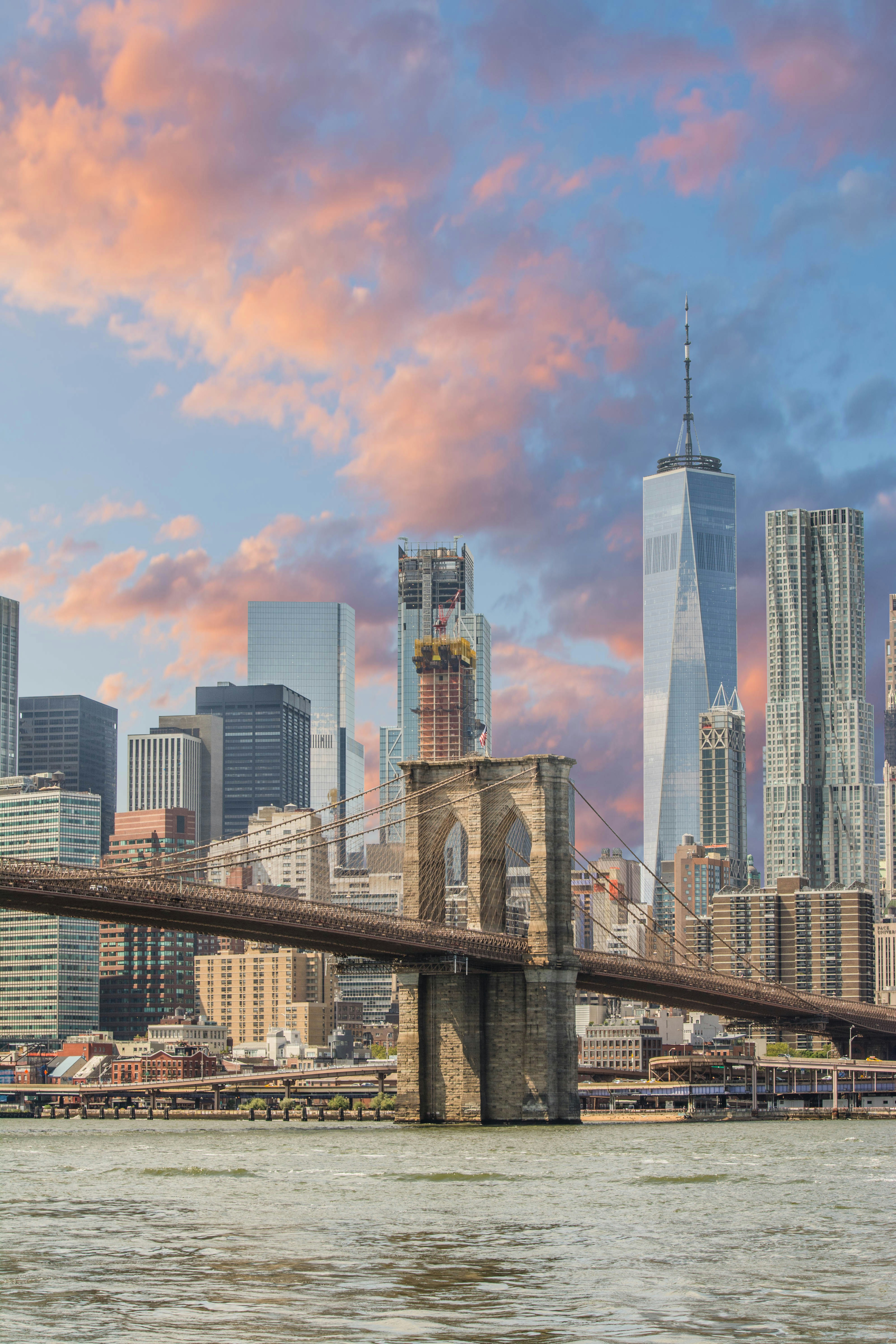 Brooklyn Bridge over water with New York City skyline and a colorful cloudy sky at sunset.