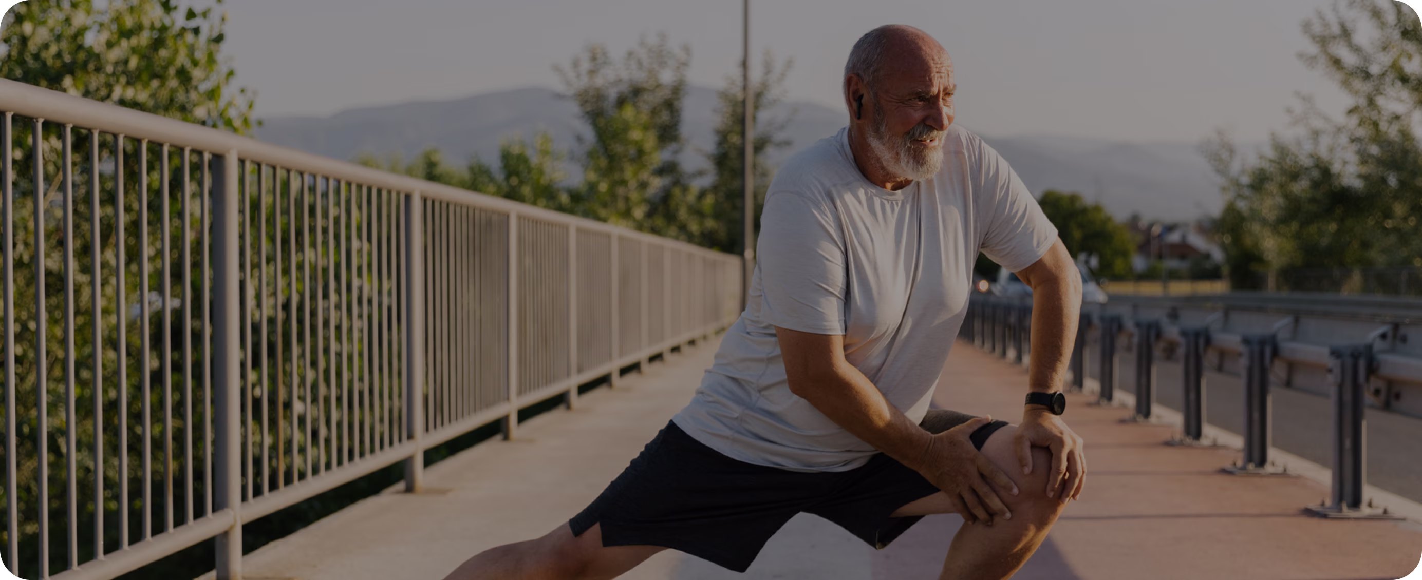 Older man with a beard stretching his legs on a bridge railing during outdoor workout.