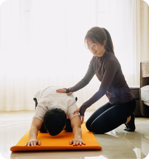 Woman assisting man doing a stretching exercise on an orange mat in a bright room.