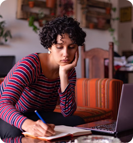 Young woman with curly hair writing in a notebook while sitting at a table with a laptop.