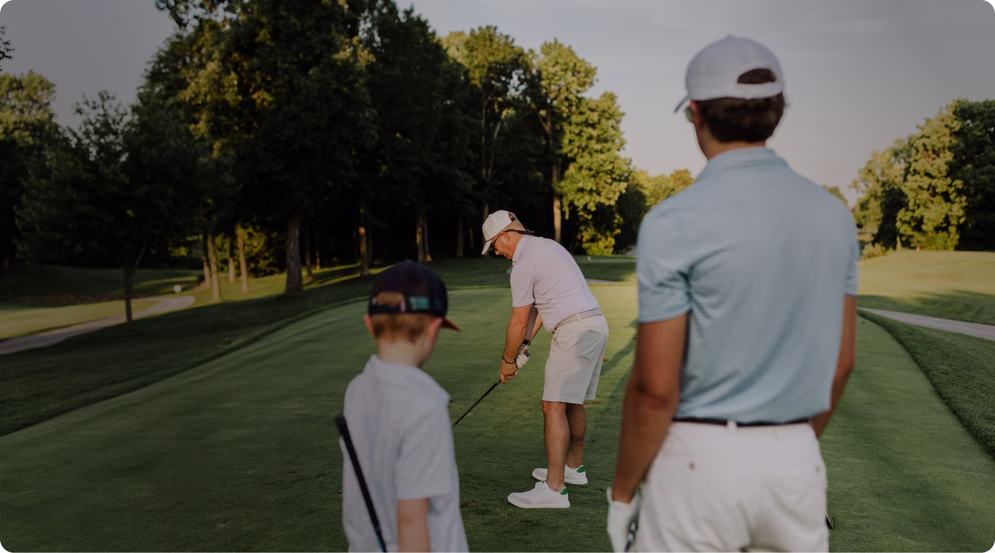 Man in light shirt and shorts preparing to swing golf club while two others watch on a green course.