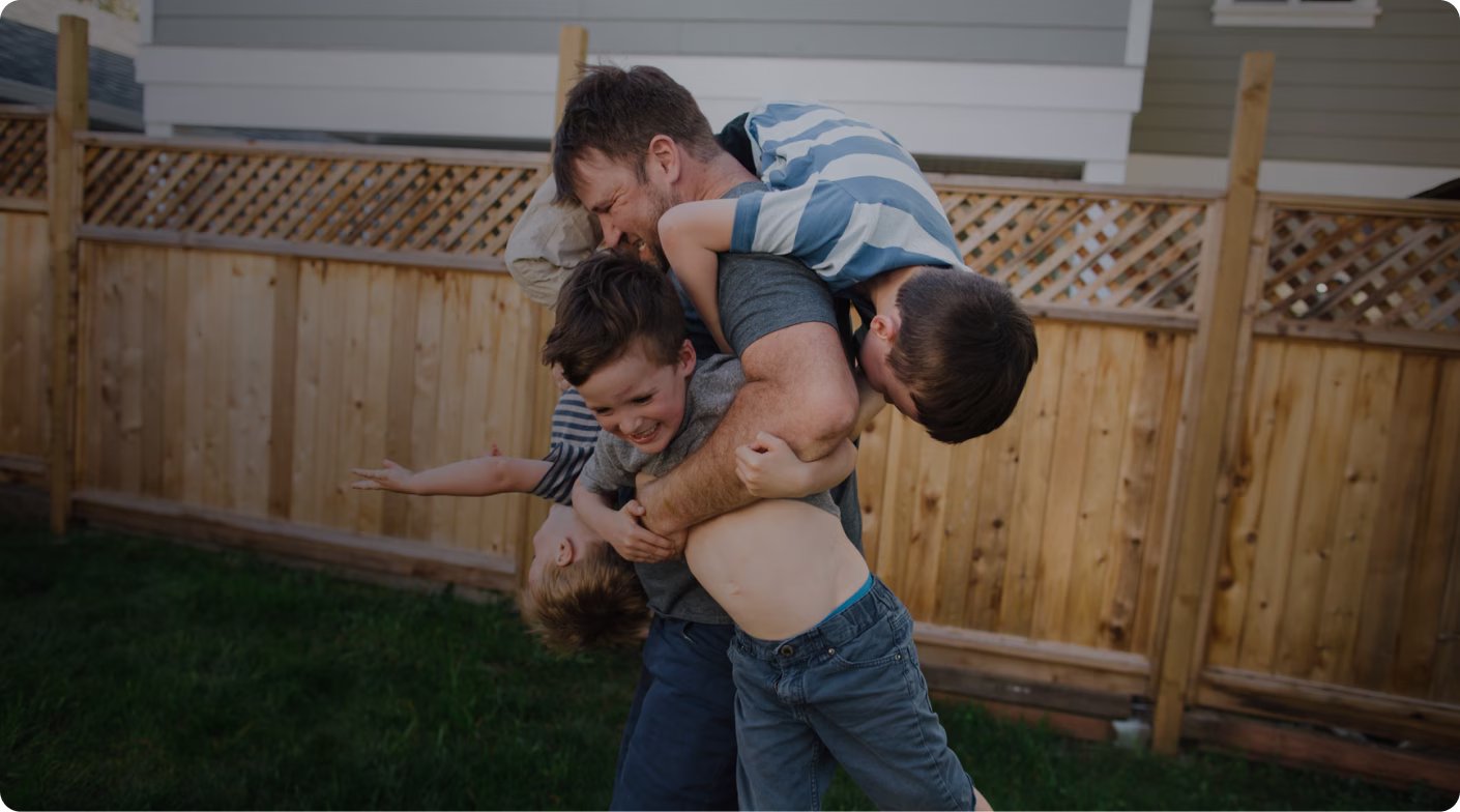 Man playfully holding and hugging three boys in a backyard with a wooden fence.