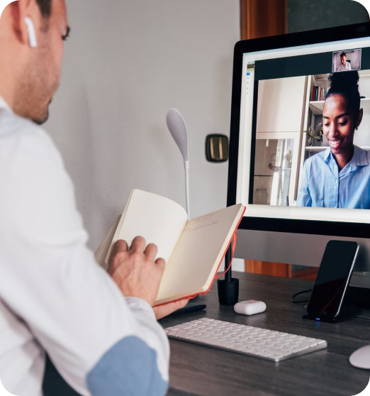 Man with earbuds reading notes during a video call with a smiling woman on a computer screen.