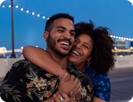 Smiling couple embracing outdoors at dusk with string lights in the background.