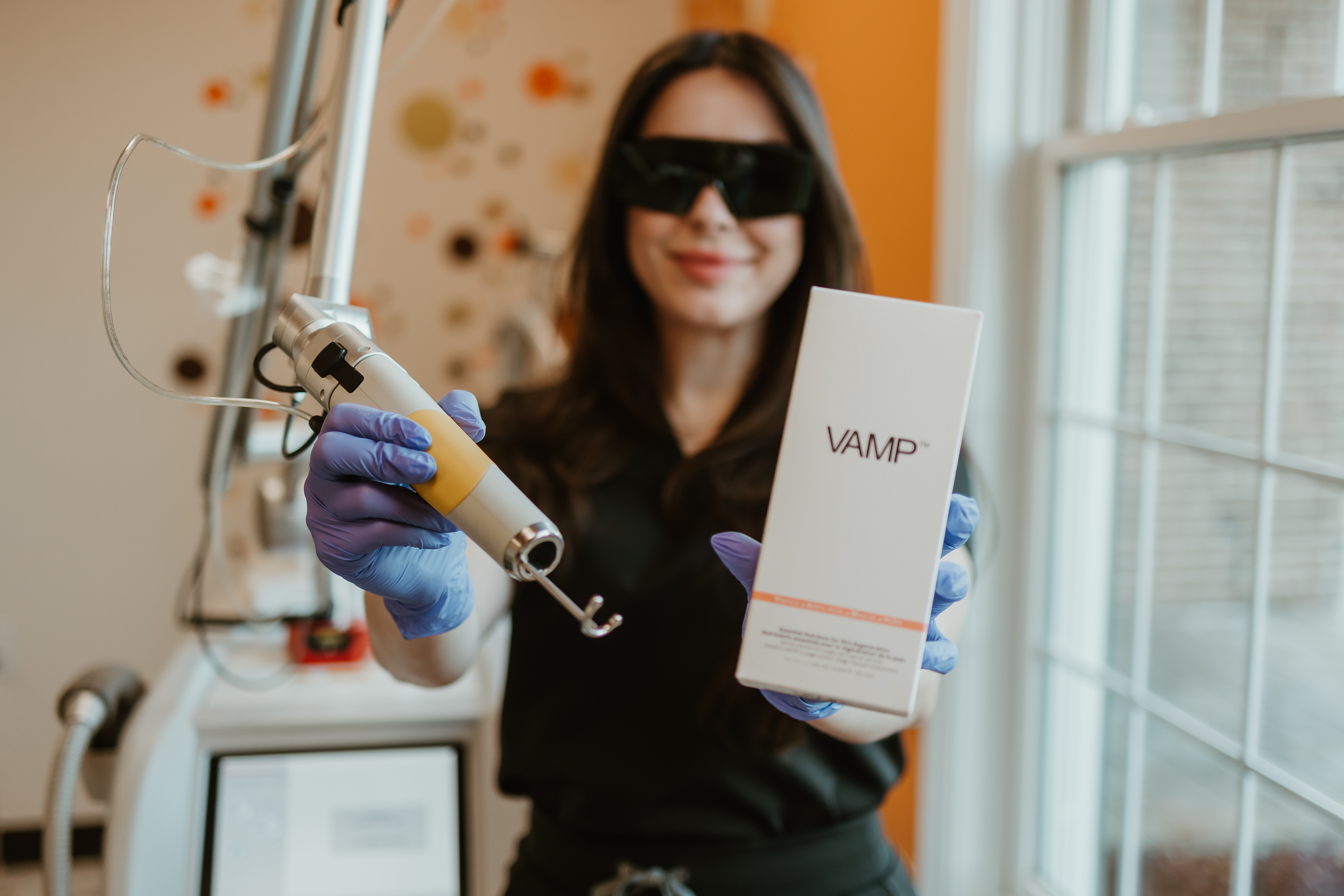 Woman wearing protective glasses and purple gloves holds a skin treatment laser device and a box labeled VAMP in a clinic room.