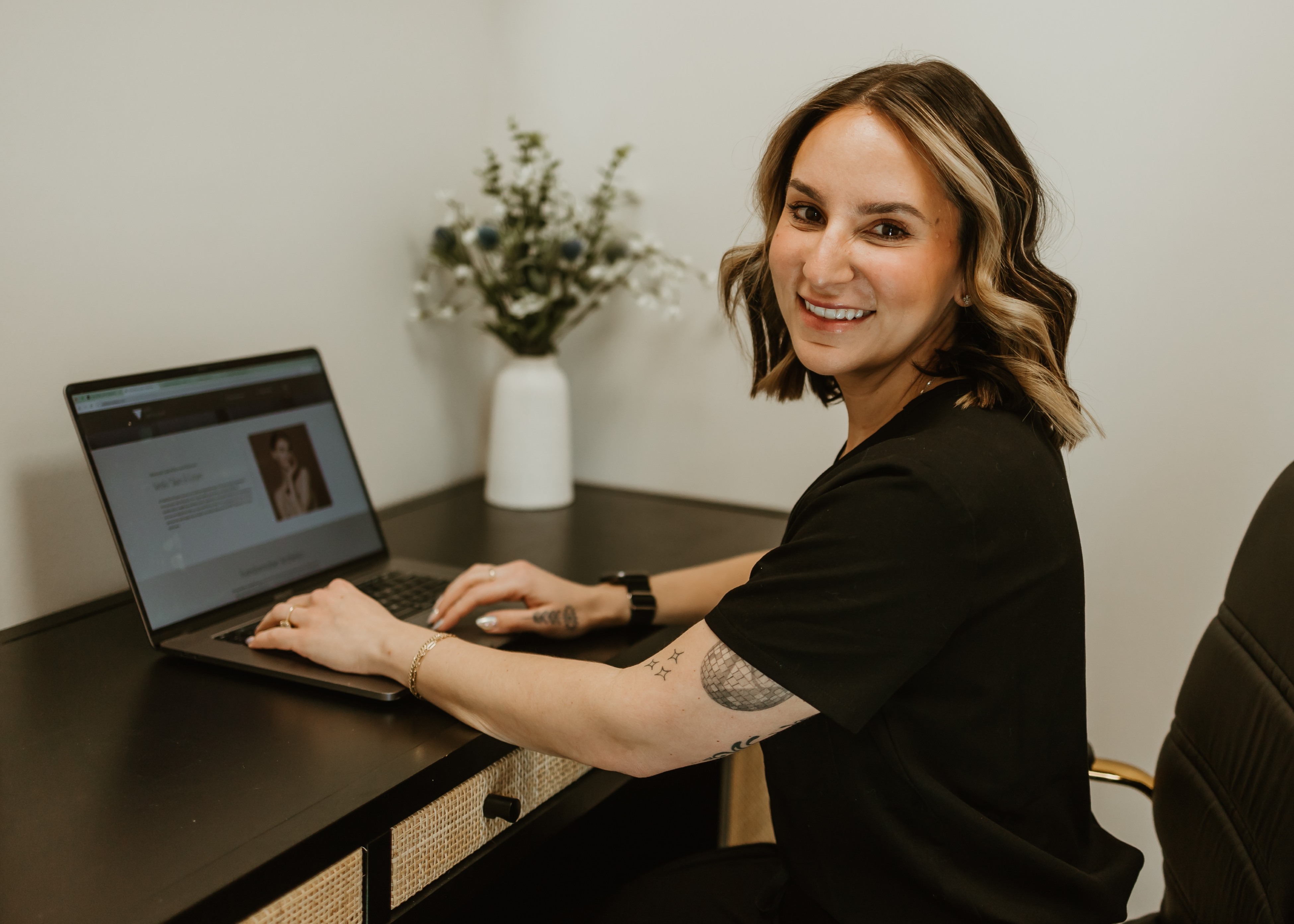 Smiling woman with tattoos sitting at a desk typing on a laptop, with a white vase and flowers in the background.