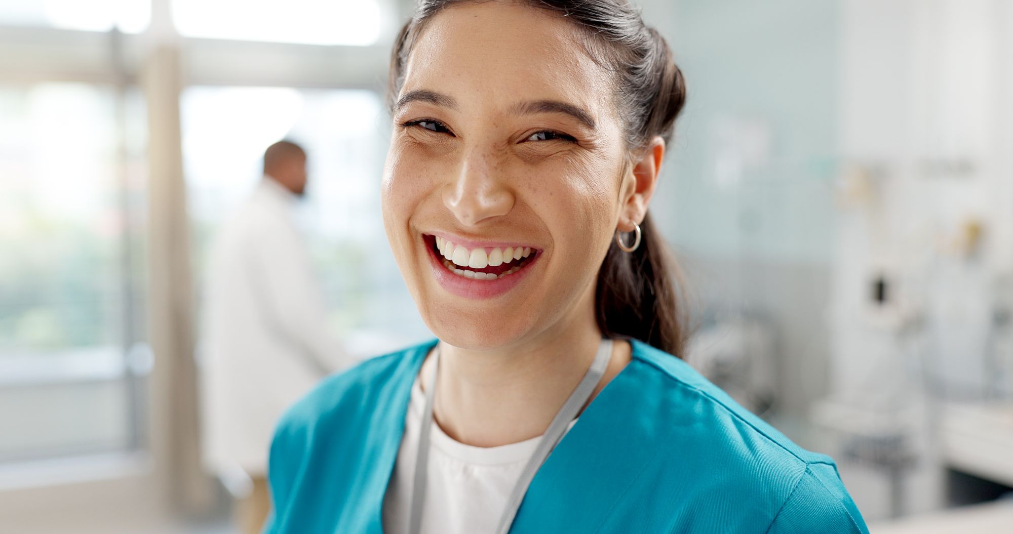 Smiling female healthcare worker in blue scrubs with a stethoscope in a medical setting.