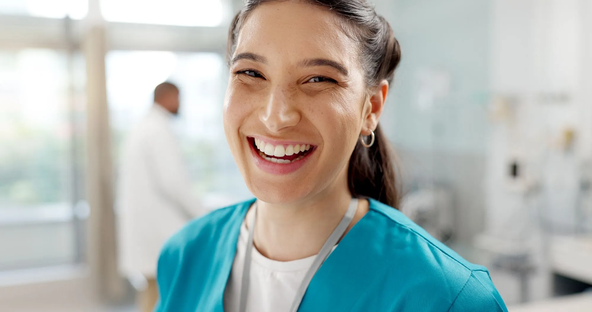 Smiling female healthcare worker in blue scrubs with a stethoscope in a medical setting.