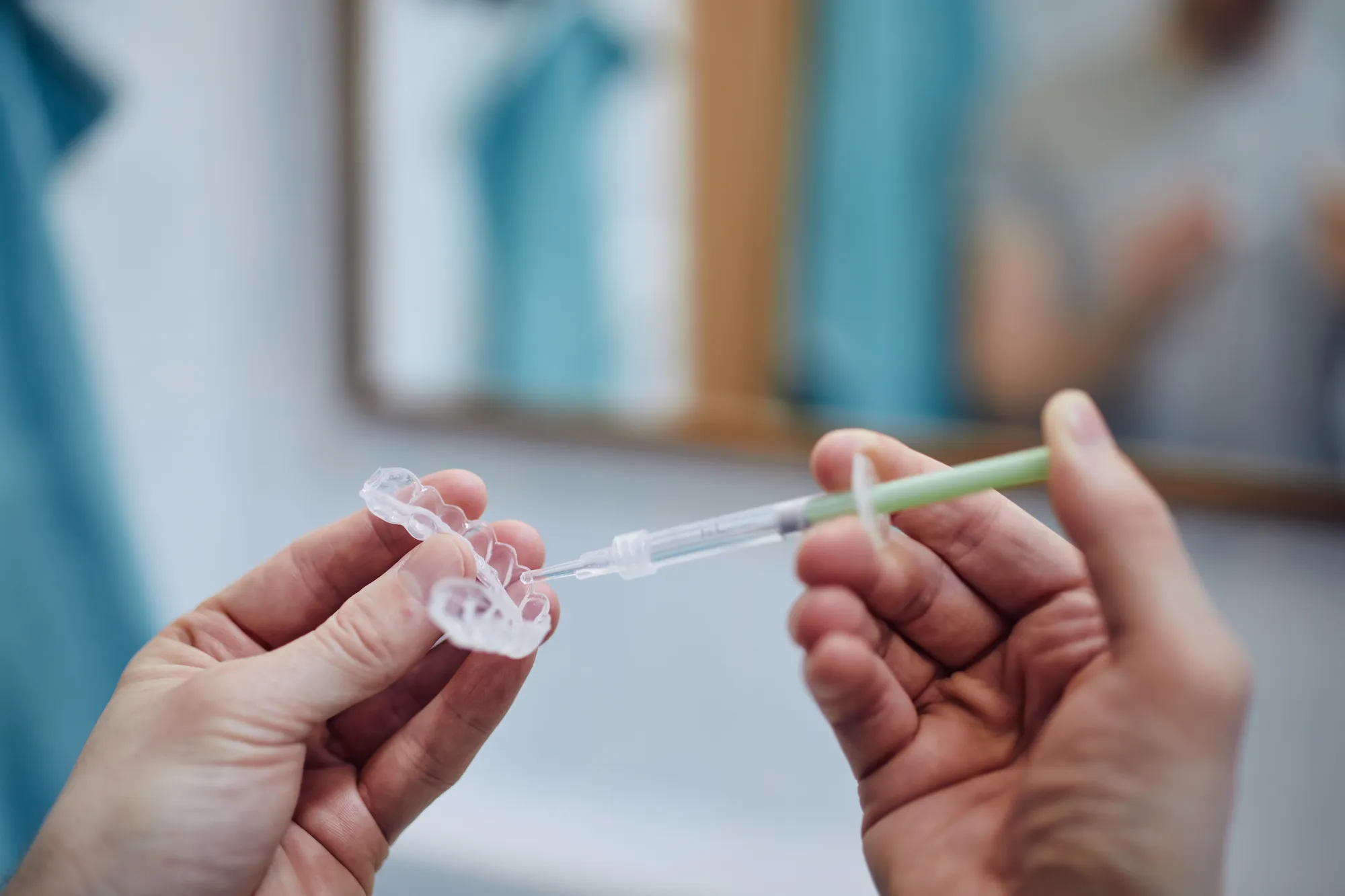 Hands applying whitening gel to a dental silicone tray using a syringe in a bathroom.